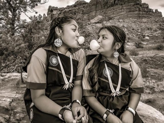 Two sisters, Acadia and Danae from Laguna Pueblo, sit on a log and look at each other while chewing gum and blowing bubbles. They are wearing traditional dress and jewelry of Laguna Pueblo. The backdrop is the beaautiful northern New Mexico landscape.