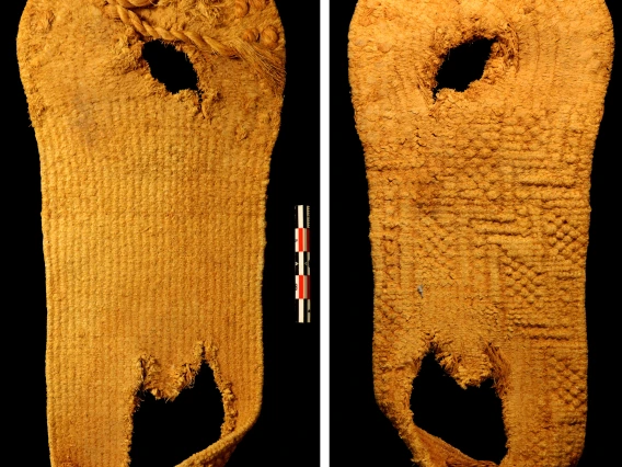 The sandal, like all the sandals in this exhibit, is made out of yucca fiber which is an ochre shade of orange. It has a round toe and a puckered heel shape. The tattered remains of a toe strap are visible. It has a large hole under the heel and a smaller hole under the ball of the foot.