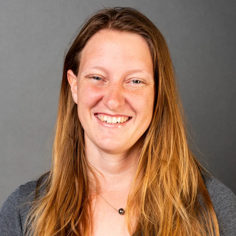 woman with long, light brown hair, wearing a gray shirt, smiles into the camera
