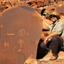 a man wearing a hat is kneeling next to a tall stone slab that shows petryglyphs on it